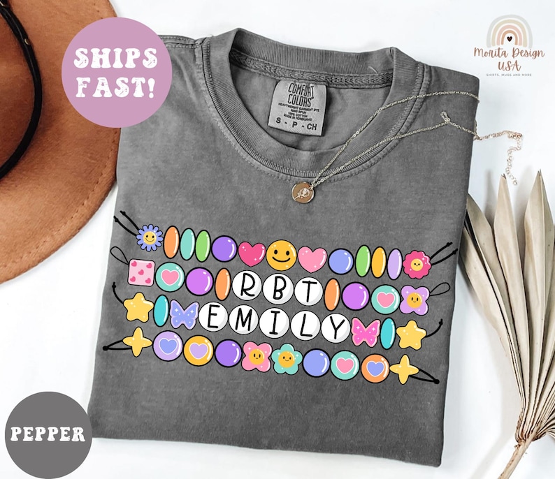 May include: A gray Comfort Colors t-shirt with a colorful beaded design that spells out "RBT EMILY". The design includes various shapes and colors, such as hearts, stars, and flowers. A gold necklace is draped across the shirt.