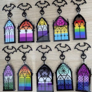 May include: Ten acrylic keychains shaped like gothic arched windows with black frames and bat-shaped clasps. Each window features a unique colourful gradient pattern representing various pride flags, including rainbow, lesbian, non-binary, and asexual designs.