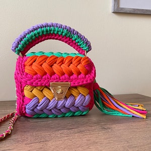 May include: A small, colorful crocheted handbag with a rainbow pattern. The bag has a top handle and a tassel. The bag is made of a variety of colors, including pink, orange, yellow, green, and purple.
