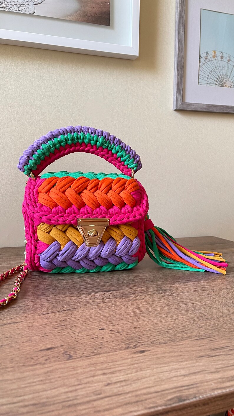 May include: A small, colorful crocheted handbag with a rainbow pattern. The bag has a top handle and a tassel. The bag is made of a variety of colors, including pink, orange, yellow, green, and purple.