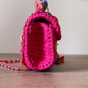 May include: A small, pink crocheted handbag with a gold chain strap. The bag has a colorful patchwork design with purple, green, yellow, and orange accents.