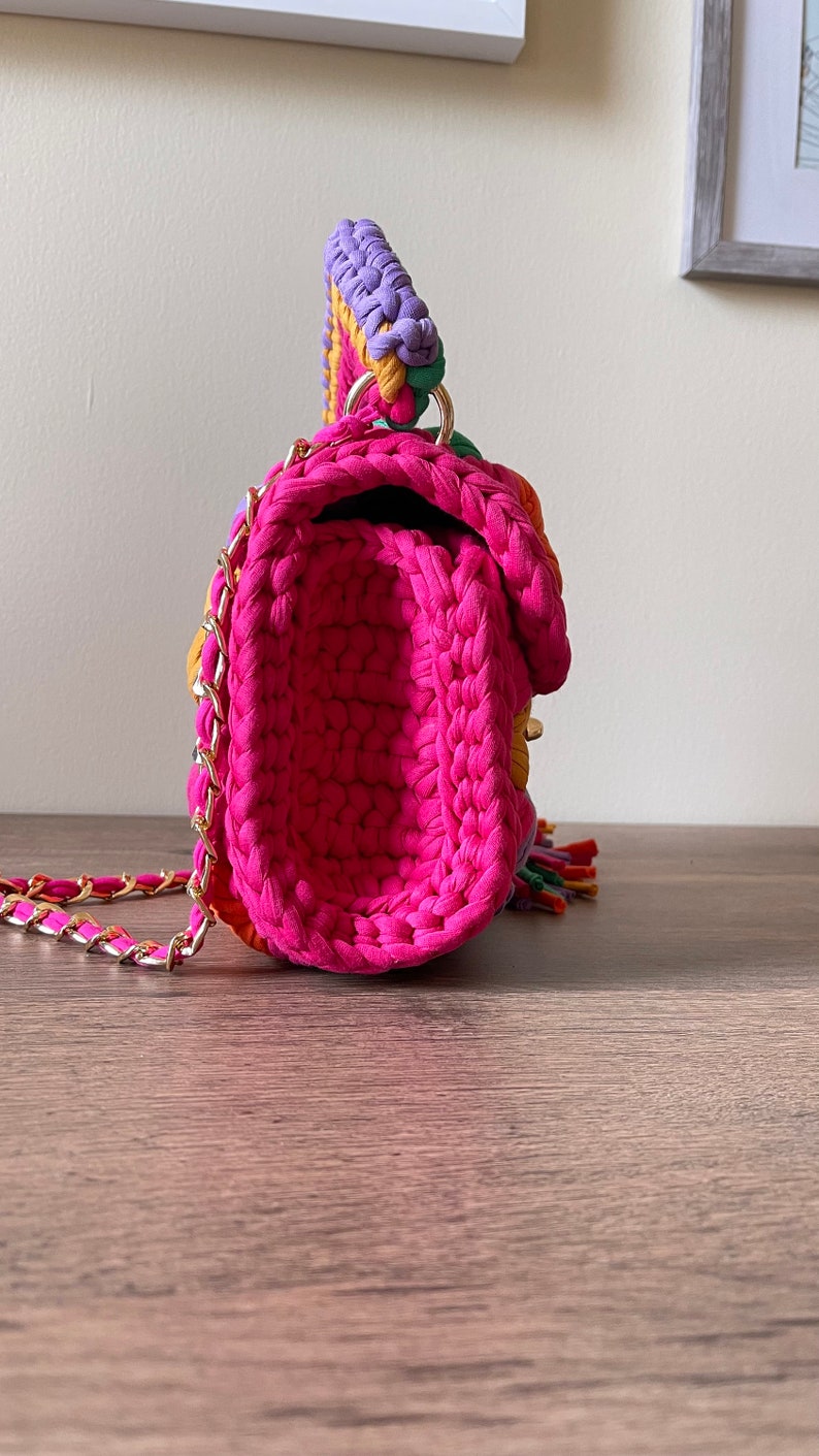 May include: A small, pink crocheted handbag with a gold chain strap. The bag has a colorful patchwork design with purple, green, yellow, and orange accents.