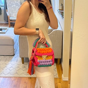 May include: A white crochet maxi dress with a side slit, worn with a colorful crocheted handbag with a gold clasp and a multi-colored tassel.