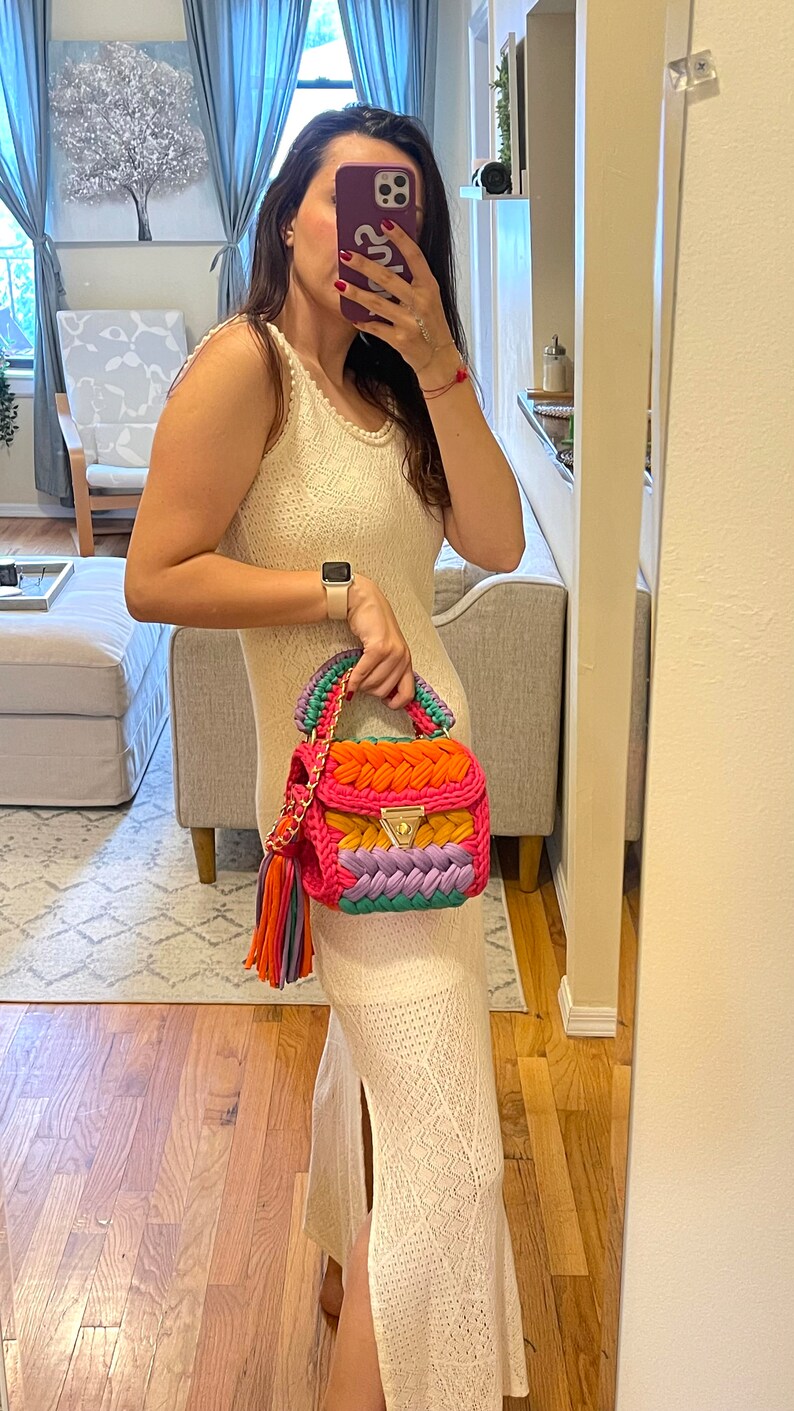 May include: A white crochet maxi dress with a side slit, worn with a colorful crocheted handbag with a gold clasp and a multi-colored tassel.