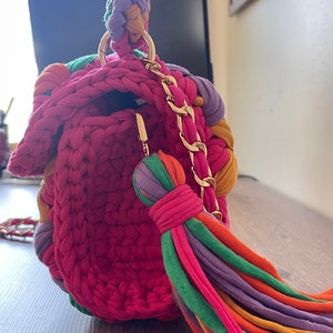 May include: A small, crocheted handbag with a colorful tassel and a gold chain strap. The bag is made of a variety of colors, including pink, purple, green, orange, and yellow.