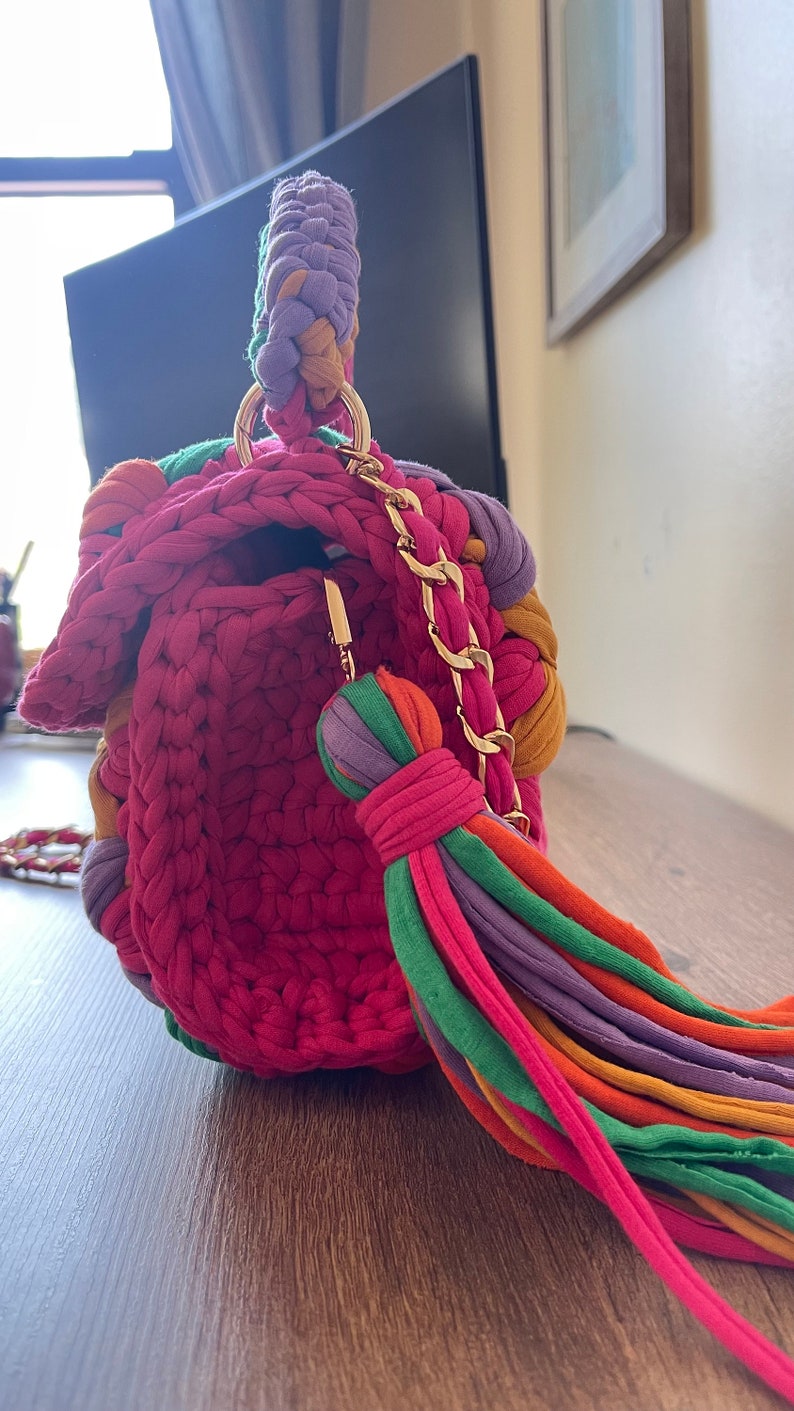 May include: A small, crocheted handbag with a colorful tassel and a gold chain strap. The bag is made of a variety of colors, including pink, purple, green, orange, and yellow.