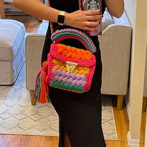 May include: A woman is holding a pink, orange, green, and purple crocheted handbag with a gold clasp and tassel. She is wearing a black sleeveless dress and a gold watch.