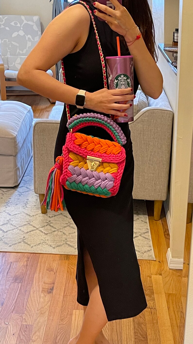 May include: A woman is holding a pink, orange, green, and purple crocheted handbag with a gold clasp and tassel. She is wearing a black sleeveless dress and a gold watch.