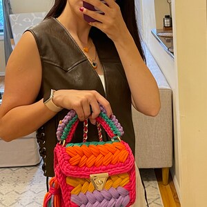 May include: A colorful crocheted handbag with a gold chain strap and a tassel. The bag is made of pink, orange, yellow, purple, and green yarn.