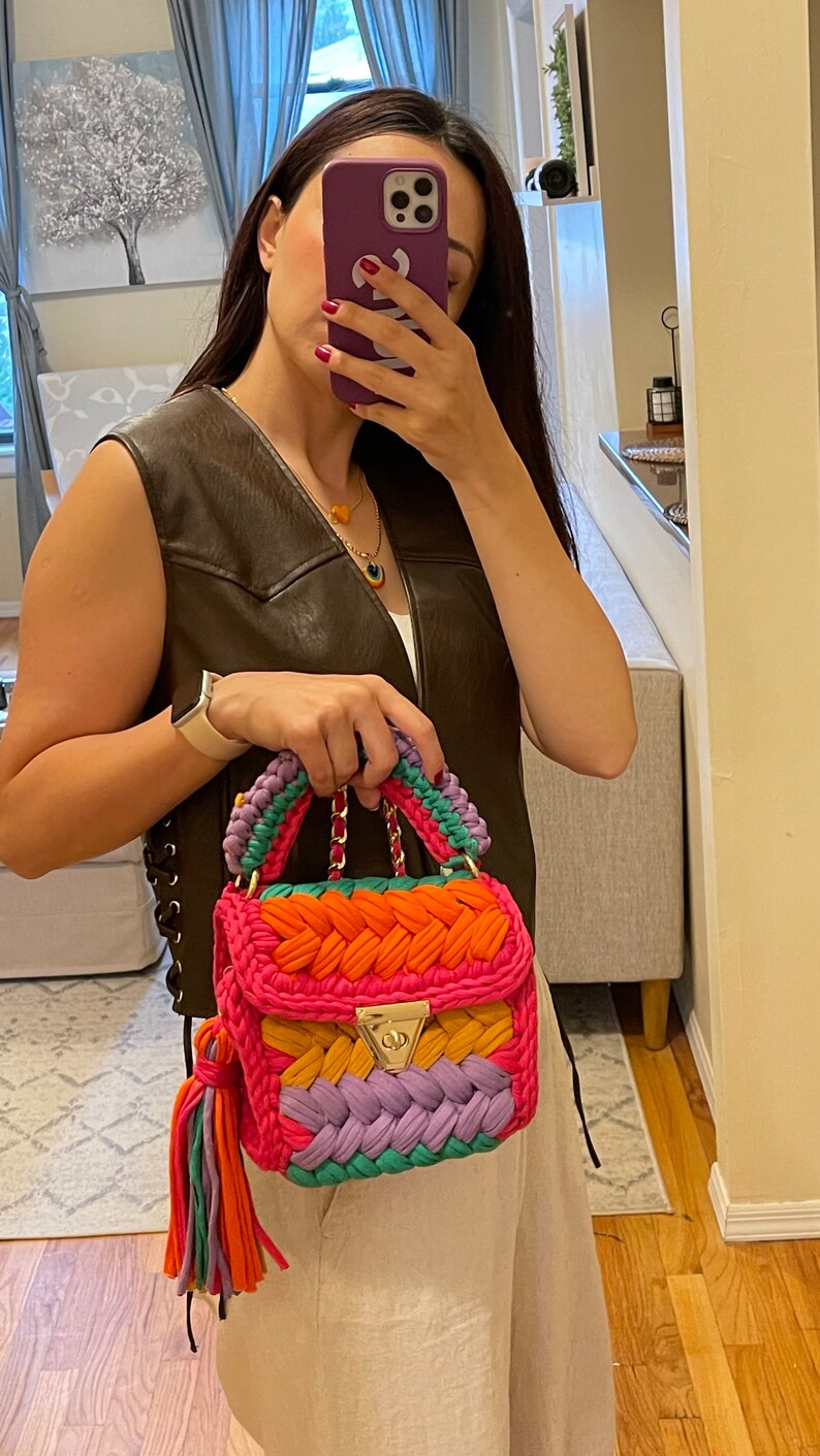 May include: A colorful crocheted handbag with a gold chain strap and a tassel. The bag is made of pink, orange, yellow, purple, and green yarn.