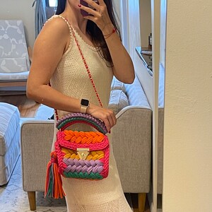 May include: A woman wearing a white crochet dress with a colorful crocheted handbag with a gold chain strap. The bag has a tassel and is made of different colored yarn.