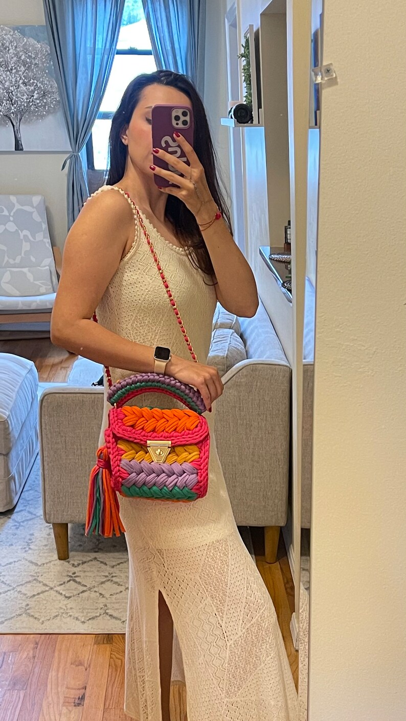 May include: A woman wearing a white crochet dress with a colorful crocheted handbag with a gold chain strap. The bag has a tassel and is made of different colored yarn.