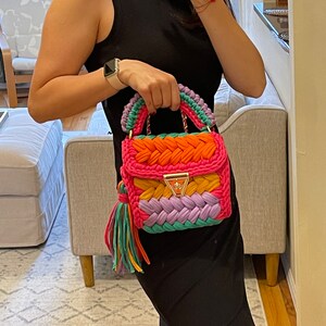 May include: A colorful crocheted handbag with a pink, orange, yellow, green, and purple design. The bag has a gold chain strap and a tassel.