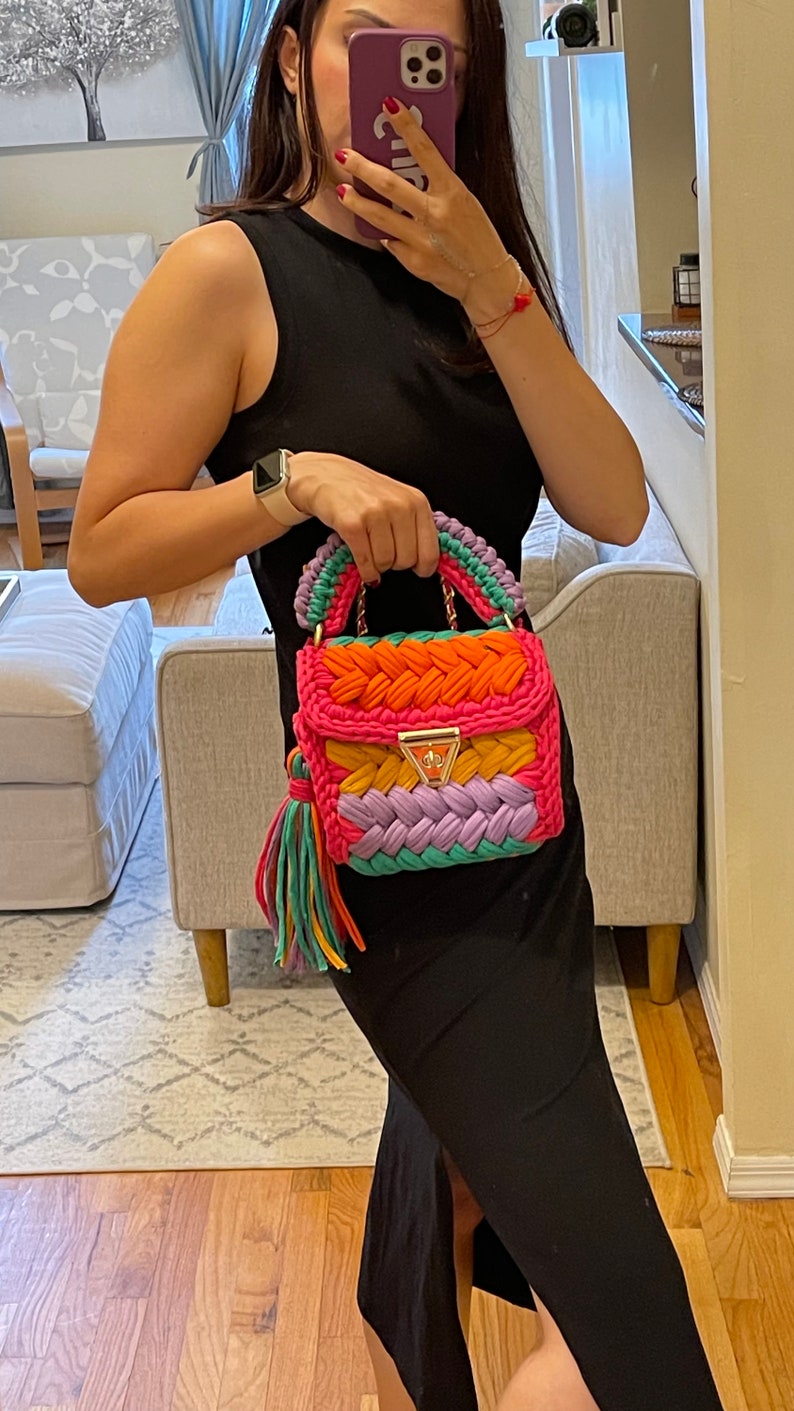 May include: A colorful crocheted handbag with a pink, orange, yellow, green, and purple design. The bag has a gold chain strap and a tassel.