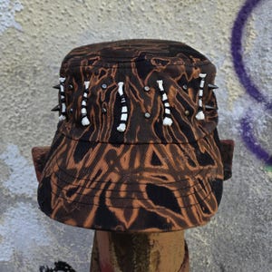 Könnte beinhalten: Eine braun-schwarze Cadet-Mütze mit Camouflage-Muster, weißen und schwarzen Akzenten und silbernen Nieten. Die Mütze hat mehrere schwarze Stacheln. Ein einzigartiges Modeaccessoire.