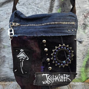 Könnte beinhalten: Dunkelblaue Denim- und Samt-Umhängetasche mit Reißverschluss. Die Tasche hat einen Pilzaufnäher, ein Zahnrad-Ornament und den Text "Jesshmäñter". Silberne Nieten und ein schwarzer Riemen vervollständigen das Design.