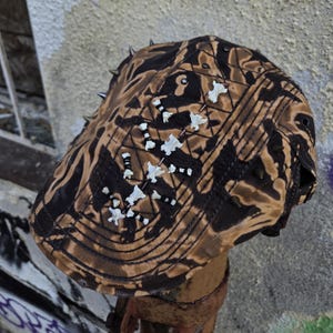 Könnte beinhalten: Eine braun-schwarz gemusterte Flatcap mit dekorativen weißen Knochenakzenten und Metallspitzen. Die Kappe hat ein einzigartiges, kantiges Design, das für alternative Mode geeignet ist.