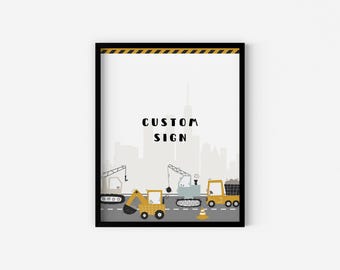 May include: A black framed print with a white background and a cityscape illustration. The print features a yellow construction truck, a yellow excavator, and a gray crane. The text "CUSTOM SIGN" is printed in black at the top of the print.