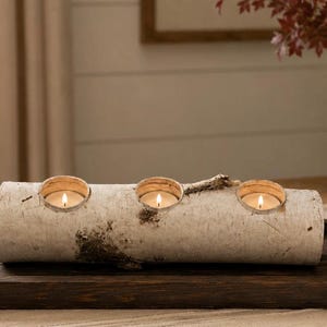 May include: A cylindrical white birch log candle holder featuring three circular cutouts, each containing a small lit tea light candle. The natural bark exterior displays textured grey and brown markings along the length of the wooden piece.
