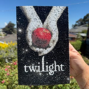May include: A black, sparkly book cover with the word "twilight" in white script. The design shows a red apple held between two white hands, all decorated with sparkling rhinestones.