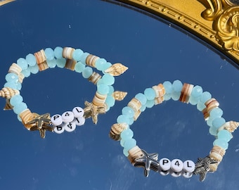 Könnte beinhalten: Zwei türkisfarbene und braune Perlenarmbänder mit silbernen Buchstabenperlen, die "P4L" buchstabieren, und goldenen und silbernen Seestern-Anhängern.