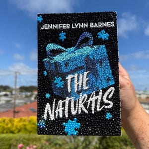 Könnte beinhalten: Ein schwarzer Buchumschlag mit dem Titel "THE NATURALS" in Weiß und dem Namen des Autors "JENNIFER LYNN BARNES" in Weiß. Der Umschlag zeigt ein blaues Geschenkbox-Design und blaue Puzzleteile.