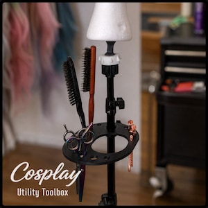 May include: A black metal stand featuring a white foam wig head topper and a circular tray with multiple slots. The tray holds two hairbrushes, one with a wooden handle, and two pairs of iridescent shears. The text Cosplay Utility Toolbox is visible.