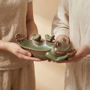May include: A ceramic frog-shaped dish in shades of green and brown, with two small bird figurines and a seashell accent. The dish is held by two people. This decorative piece is ideal for holding small items.