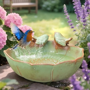 May include: A ceramic bird bath with a light green finish, featuring a decorative leaf design and two small bird figurines. A blue and orange bird is shown mid-flight, splashing water. The bird bath is surrounded by pink and purple flowers.