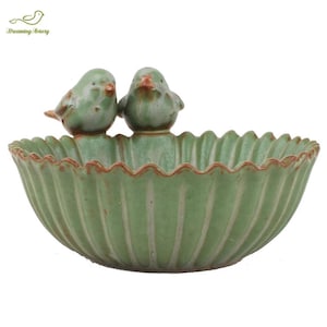 May include: A green ceramic bowl with a scalloped rim and vertical grooves. Two small green bird figurines perch on the rim. The bowl has a rustic, aged appearance, with brown accents. The product is likely a decorative item or a small serving dish.