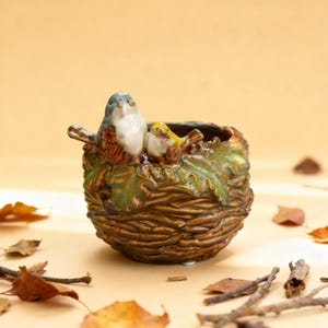 May include: A ceramic planter shaped like a bird's nest, featuring two decorative birds perched on the rim. The planter is brown with green leaf accents, and the birds are painted in shades of brown, white, and yellow. A charming decorative item.