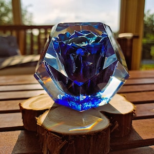 May include: A blue rose encased in a clear, faceted resin block. The floral arrangement sits atop three small, round wooden slices. The background features a wooden table and a blurred outdoor setting.