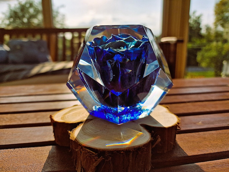 May include: A blue rose encased in a clear, faceted resin block. The floral arrangement sits atop three small, round wooden slices. The background features a wooden table and a blurred outdoor setting.