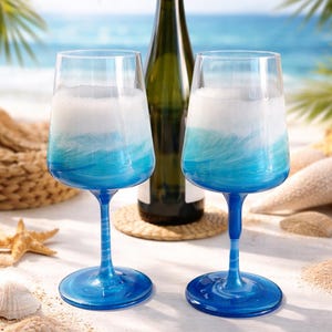 Könnte beinhalten: Zwei Weingläser mit blauem Wellendesign, gefüllt mit einer weißen Flüssigkeit, stehen auf einer weißen Oberfläche mit Sand und Muscheln. Im Hintergrund steht eine grüne Weinflasche. Die Gläser haben blaue Stiele und Böden.