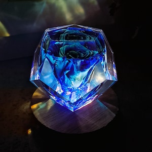 May include: A blue rose encased in a clear, faceted resin block. The block sits on a round wooden base. The rose is a deep blue color, and the resin reflects light, creating a glowing effect.