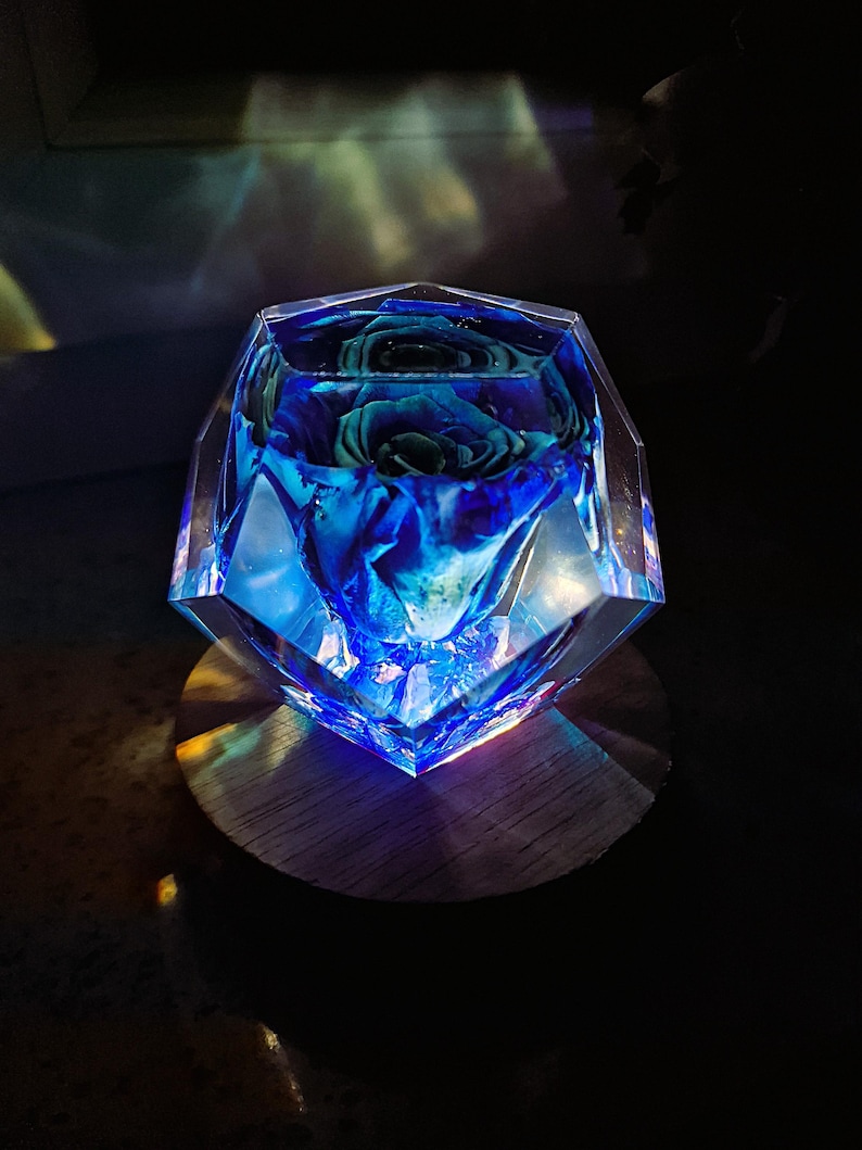 May include: A blue rose encased in a clear, faceted resin block. The block sits on a round wooden base. The rose is a deep blue color, and the resin reflects light, creating a glowing effect.