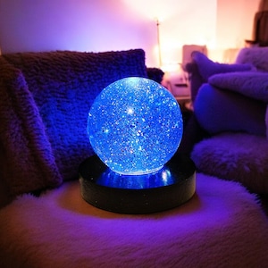 Puede incluir: Un globo de vidrio azul con purpurina en su interior se asienta sobre una base de madera negra. El globo está iluminado desde el interior, proyectando un brillo azul.