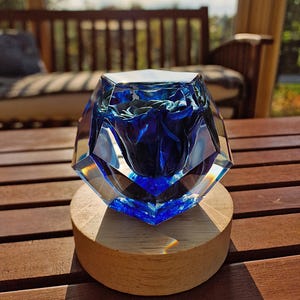 May include: A faceted, clear glass sculpture with a deep blue interior, resting on a round wooden base. The sculpture's geometric design catches the light, creating a play of colors. The wooden base is approximately 3 inches in diameter.