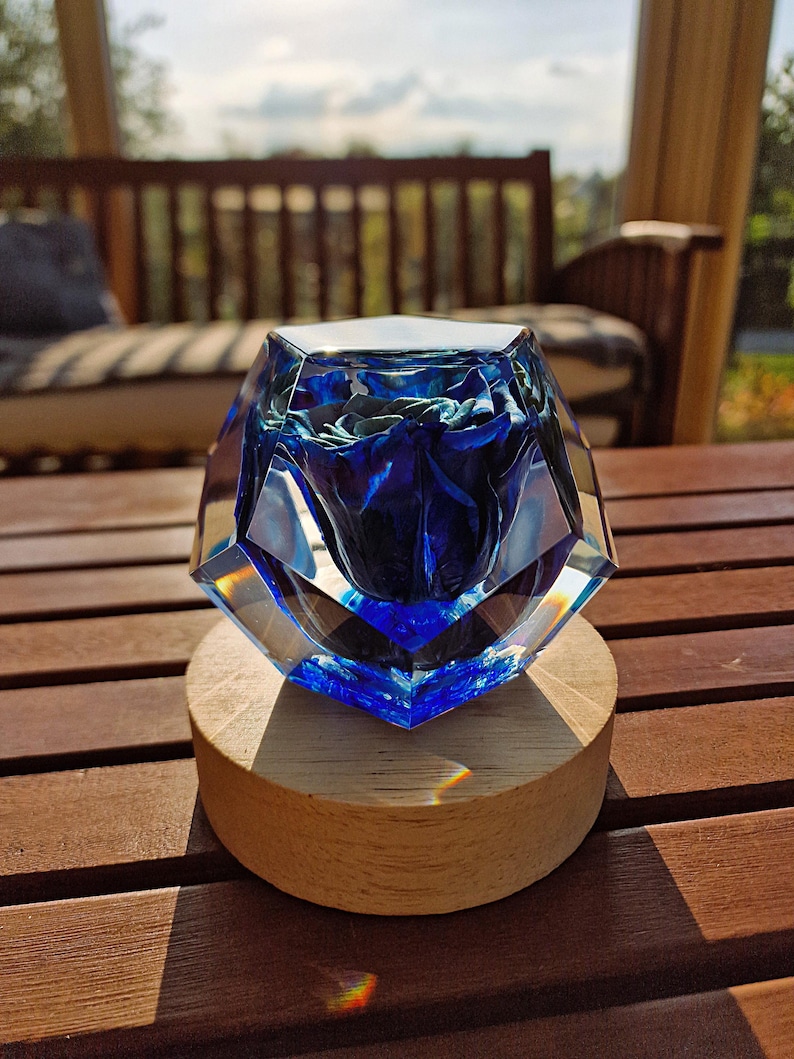 May include: A faceted, clear glass sculpture with a deep blue interior, resting on a round wooden base. The sculpture's geometric design catches the light, creating a play of colors. The wooden base is approximately 3 inches in diameter.
