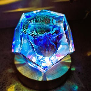 May include: A blue and clear resin art piece with a preserved blue rose inside. The geometric shape sits on a round wooden base. The light refracts through the resin, creating a colorful display.