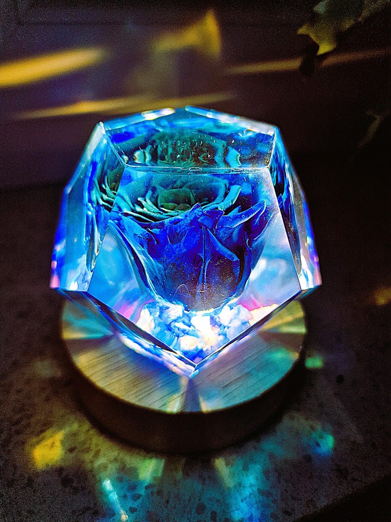 May include: A blue and clear resin art piece with a preserved blue rose inside. The geometric shape sits on a round wooden base. The light refracts through the resin, creating a colorful display.