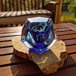 May include: A blue rose encased in a faceted, clear glass sculpture. The rose is a deep blue hue, and the glass has a geometric design. The sculpture sits on a natural wood slice, displayed on a wooden table.