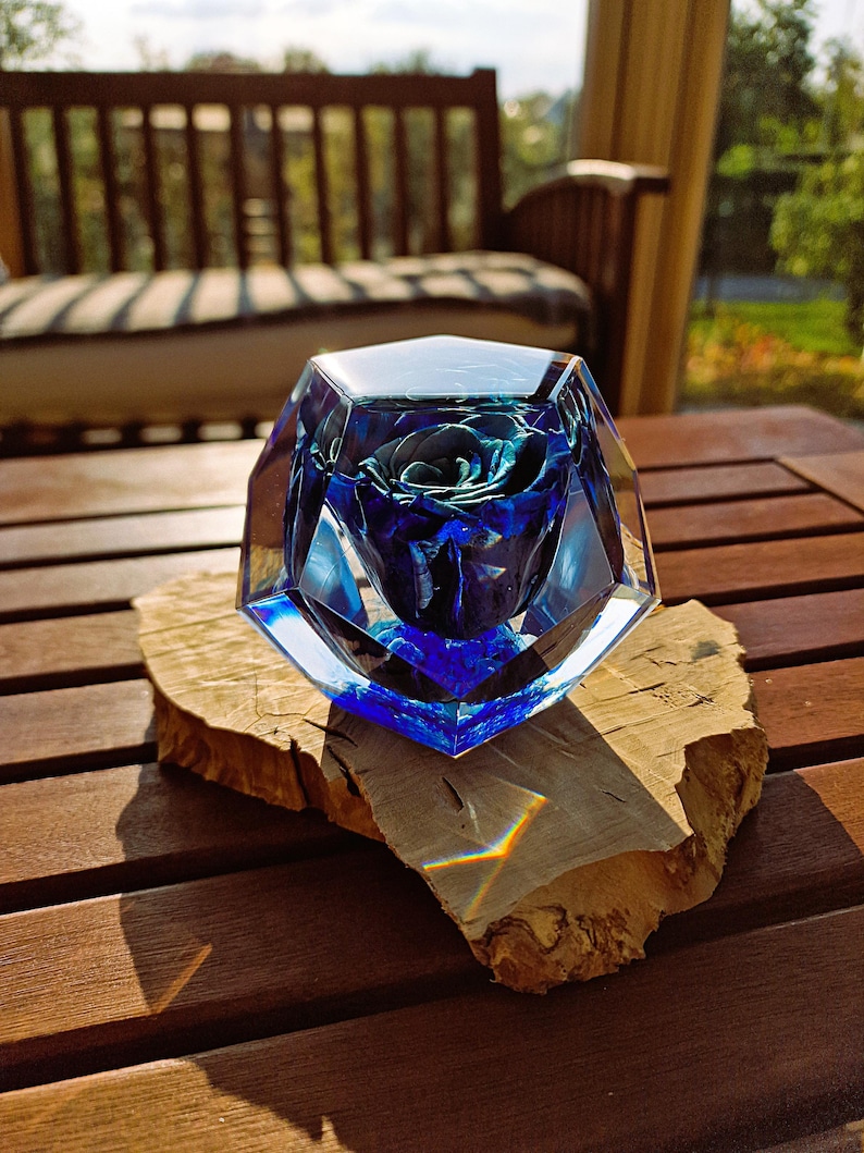 May include: A blue rose encased in a faceted, clear glass sculpture. The rose is a deep blue hue, and the glass has a geometric design. The sculpture sits on a natural wood slice, displayed on a wooden table.