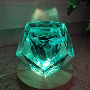 May include: A teal-colored, faceted resin sculpture with a preserved dark rose inside. The sculpture sits on a round, light-colored wooden base. The light shines through the resin, creating a glowing effect.