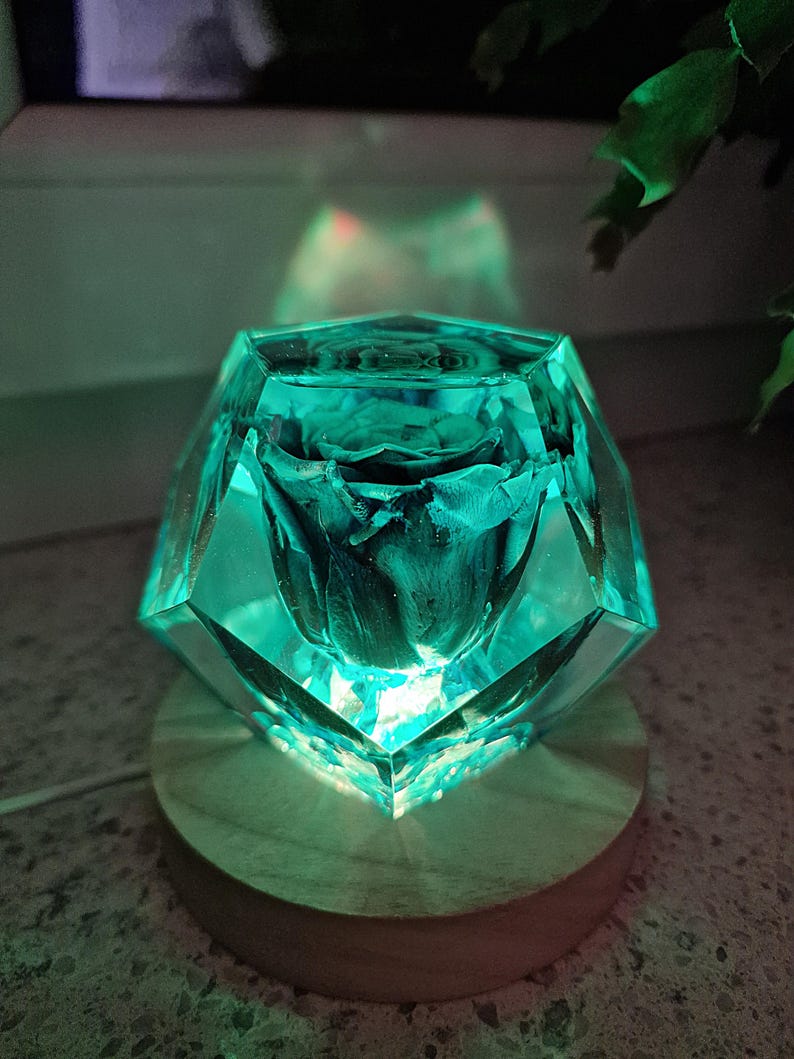 May include: A teal-colored, faceted resin sculpture with a preserved dark rose inside. The sculpture sits on a round, light-colored wooden base. The light shines through the resin, creating a glowing effect.