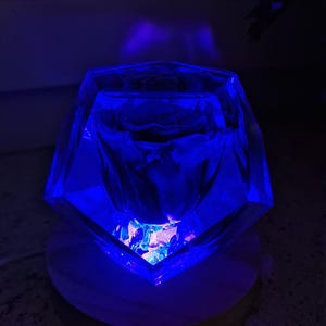 May include: A faceted, clear glass object illuminated with blue light, resting on a round, wooden base. The glass appears to be filled with smaller, colorful pieces. The lighting creates a dramatic effect, highlighting the object's geometric shape.