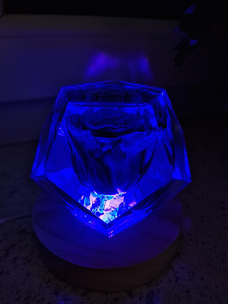 May include: A faceted, clear glass object illuminated with blue light, resting on a round, wooden base. The glass appears to be filled with smaller, colorful pieces. The lighting creates a dramatic effect, highlighting the object's geometric shape.