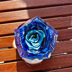 May include: A blue and clear resin art piece with a preserved blue rose inside. The resin is shaped in a geometric form and sits on a small wooden slice. The background is a wooden surface.