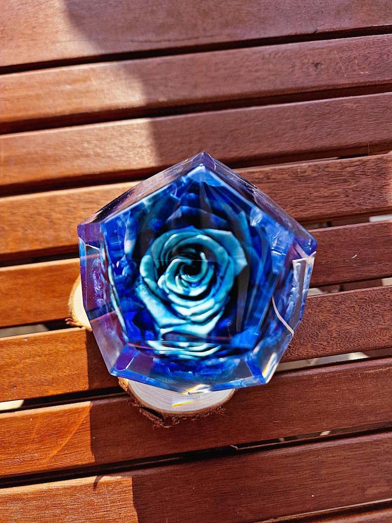 May include: A blue and clear resin art piece with a preserved blue rose inside. The resin is shaped in a geometric form and sits on a small wooden slice. The background is a wooden surface.