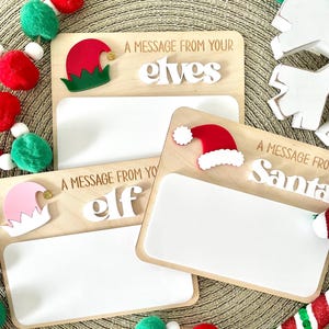 May include: Wooden message boards with the text "A Message From Your Elves" and "A Message From Santa". Each board has a white space for writing and is decorated with elf and Santa hats. Christmas-themed decor surrounds the boards.